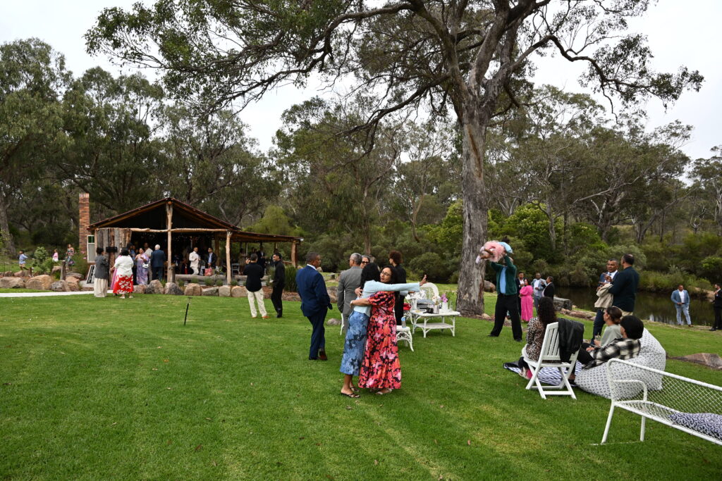 gathering of people on green lawn, some hugging each other, children playing and other guests seated chatting to one another, trees and a timber structure in background